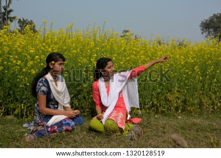Close up two beautiful girls sitting in a rapeseed, mustard flower field with two flowers on their ears smiling, talking and pointing  something, conceptual picture, selective focusing