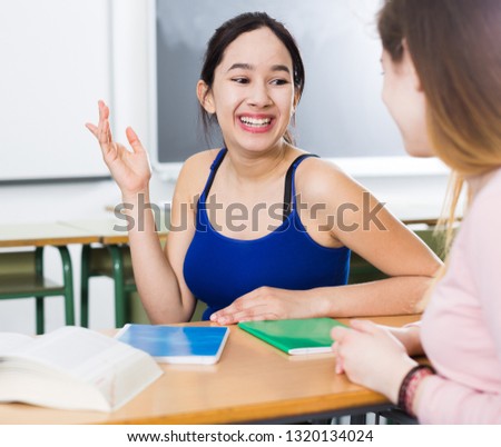 Two girl are sitting at the desk and talking about life in the classroom.