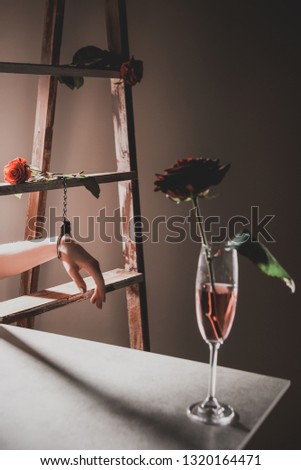 red rose flowers in champagne glass on stone table with wooden ladder and woman in metal handcuffs on background