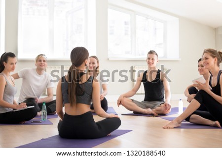 Smiling sporty men and women take part in yoga seminar, sit on mats write down in notebook listen to trainer, happy millennial people study fitness coaching at gym or training studio with teacher Royalty-Free Stock Photo #1320188540