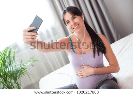 Young brunette woman sitting on the bed in her room, listening to the music in earphones and making selfie.