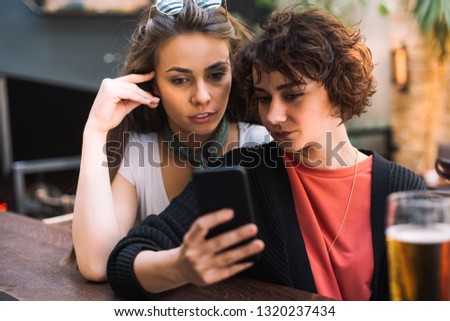 Two girls having a drink while chatting. Girls at a bar having a drink. Grls drinking beer at a bar. Girls having a beer and taking selfies.
