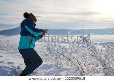 Woman taking a picture of the view with a mobile phone. Field and forest, mountains covered with snow. Blue sky. Frosty day.