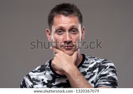 Portrait of expressive uncertain man looking at camera with hands on chin over gray background. 