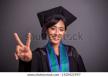 Pretty smiling Asian female student dressed for her graduation day showing victory hand sign to camera, closeup portrait on gray background