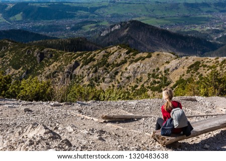 Woman is sitting on a wooden bench and enjoying a great view of mountain landscape. Summer day with blue sky and white clouds. Tourism concept. Landscape composition.