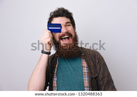 Photo of emotional excited young bearded man standing isolated over red background and covered eye with credit card 