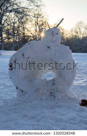 Large snow man with a hole in its belly, with tree branches on its head, standing in the snow with the sun setting in the background