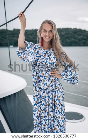 portrait of happy pretty girl with colorful dress and long curly blonde hair standing on yacht at summertime. looking at camera with toothy smile.