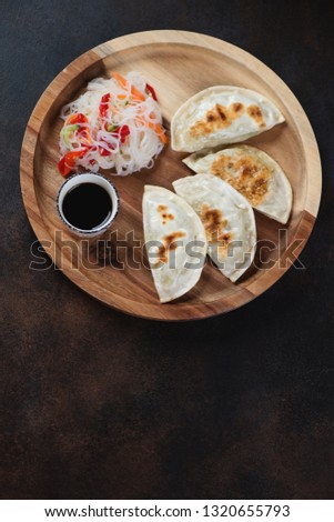 Wooden serving tray with fried potstickers, funchoza noodles salad and soy sauce. Flatlay on a dark brown metal surface with space