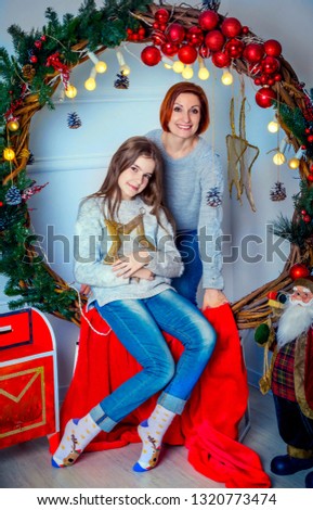 Beautiful happy smiling mother and daughter, wearing casual gray jumpers and blue jeans, posing near Christmas wreath decorated with spruce branches, red balls and fairy lights. New year portrait.