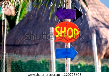 Hand made speech bubble shape shop sign and direction arrows on a post in a tropical resort with thatched huts in the background.