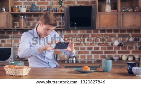 Handsome caucasian businessman taking photo his breakfast. Smiling man using smart phone using application shared picture in social media. Happy man wearing elegant suit in studio apartment. Morning