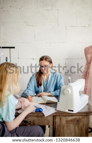 Cute female seamstress discusses with the client sketch of custom made lady s suit. Young female tailor and cloth designer collaborating in the workshop.