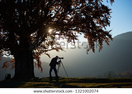 Tourist photographer taking picture of misty mountain landscape using professional camera on tripod on quiet autumn evening standing on grassy valley under large tree under blue sky at sunset.