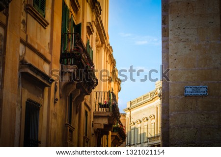 
The picture shows a beautiful old yellow building with a green balcony, built in a certain style. This street is located on the island of Malta in a city called Valletta