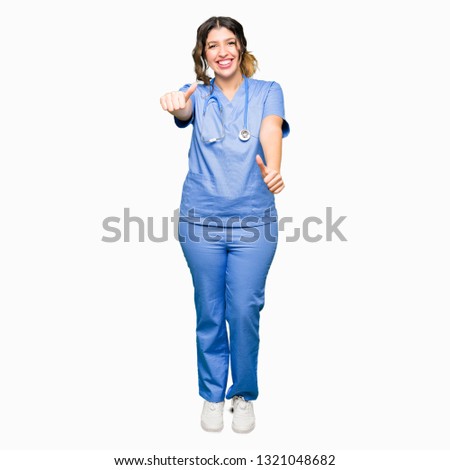 Young adult doctor woman wearing medical uniform approving doing positive gesture with hand, thumbs up smiling and happy for success. Looking at the camera, winner gesture.