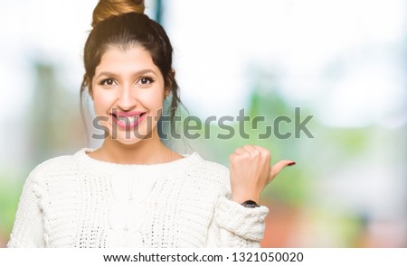 Young beautiful woman wearing winter sweater smiling with happy face looking and pointing to the side with thumb up.