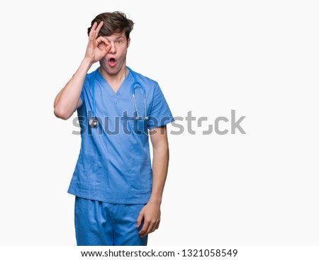 Young doctor wearing medical uniform over isolated background doing ok gesture shocked with surprised face, eye looking through fingers. Unbelieving expression.