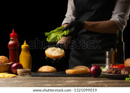 The chef puts fresh salad on a burger loaf, with the background ingredients. Delicious and harmful food, fast food, homemade recipes, restaurant, catering, recipe book