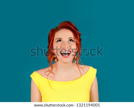 Thoughtful woman looking up happy smiling laughing showing teeth. Caucasian person in yellow dress and beige lipstick, redhead hair up isolated on blue studio background. Expressive girl human emotion