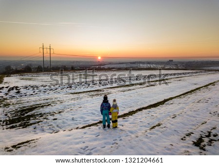 Back view of two young children in warm clothing standing in frozen snow field holding hands on copy space background of setting sun and clear blue sky.