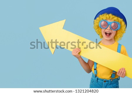 Child, sale, spring. Child in yellow,  blue colors, funny glasses, with a large  arrow directional, free space. Advertisement. 
Waist up, thin, facing camera, studio, blue background.
