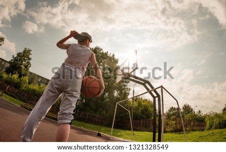 A fisheye view of a Caucasian teenager standing on the play ground with a basket-ball in his hand, under setting summer sun, getting ready for streetball training, curved tilted horizon, toned image.