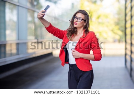 Long hair beautiful business woman in red suit with mobile phone taking selfie in front of office