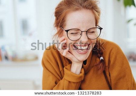 Pretty red-haired girl with pigtail, wearing glasses and orange sweatshirt, laughing with her eyes closed. Close-up front portrait indoors with copy space Royalty-Free Stock Photo #1321378283