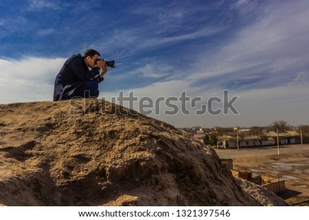 a photographer in a dark outfit and black sunglasses holding a camera steady in his hands and making a shot while sitting on top of an ancient wall. Aerial view.