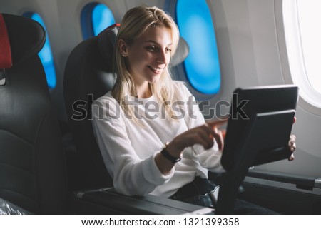 Smiling attractive female passenger searching movie to watch on board tv device during comfortable flight. Caucasian young woman traveler enjoying touch screen entertainment monitor system on airplane Royalty-Free Stock Photo #1321399358