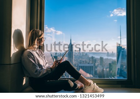 Smiling hipster girl enjoying beautiful views of New York city while sitting in hotel room on high floor of skyscraper. Happy female tourist using touch pad for communication during vacation holidays