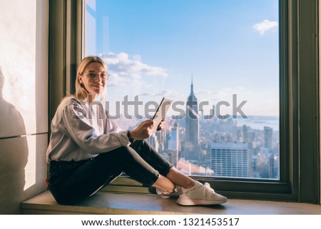 Successful Caucasian female freelancer enjoying convenience of remote work while traveling to United States. Happy hipster girl traveler sitting on window sill with touch pad in hands and smiling