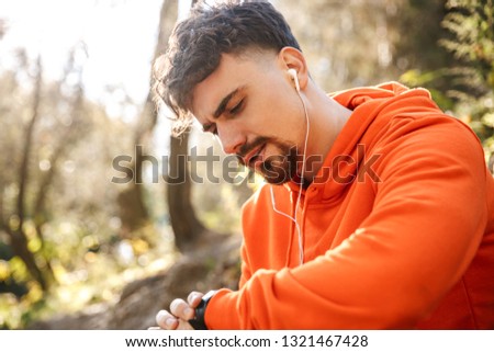 Picture of handsome young sports fitness man runner outdoors in park listening music with earphones looking at watch clock.