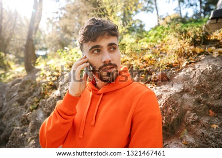 Picture of handsome serious young sports fitness man runner outdoors in park talking by mobile phone.