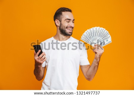 Photo of smiling man 30s in casual wear holding cell phone and fan of money isolated over yellow background
