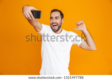 Photo of cheerful guy 30s in casual wear screaming and taking selfie on cell phone isolated over yellow background