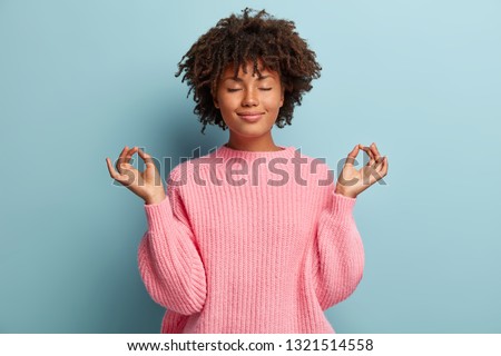 People, peace and meditation concept. Calm black young woman practices yoga indoor, shows okay sign with both hands, demonstrates approval, wears pink clothing, isolated over blue background Royalty-Free Stock Photo #1321514558