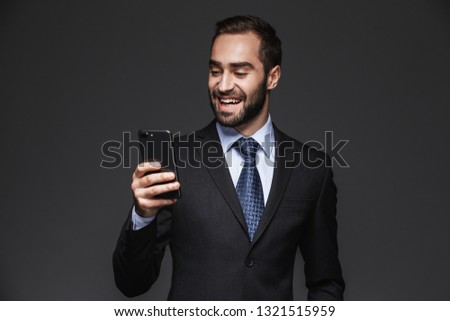 Portrait of a confident handsome businessman wearing a suit standing isolated over black background, using mobile phone