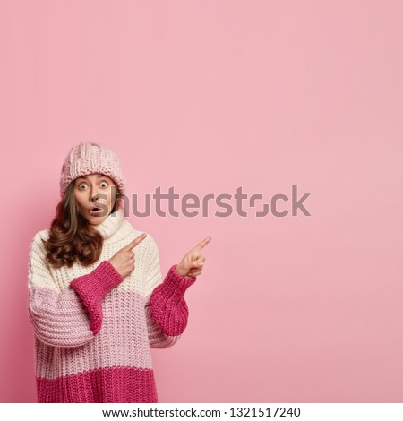 Vertical shot of surprised woman with dark curly hair, being in stupor, demonstrates big sales in shop, wears headgear, oversized jumper, points with both fore fingers at blank space, isolated on pink