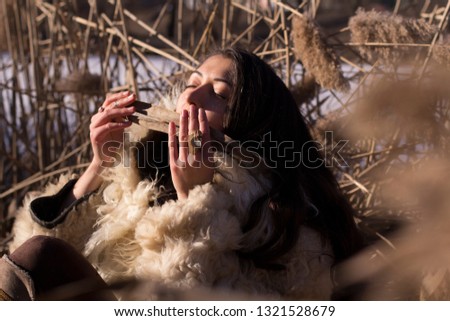 Young brown-haired charming beautiful girl with long hair in fur coat in swamp with reeds sees off the winter and meets spring near ice lake