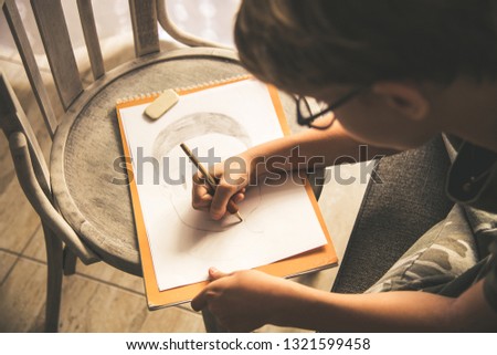 Close up top view of a young boy drawing a face on a white sheet. Kid hold a pencil and draw something, warm orange light at home. Children writing on a paper. Teen drawing freehand a manga head.