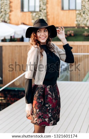 Stylish girl dressed in a black turtleneck, beige cape, stylish short skirt and black hat with wide bells poses in the street in the city on the sunny day