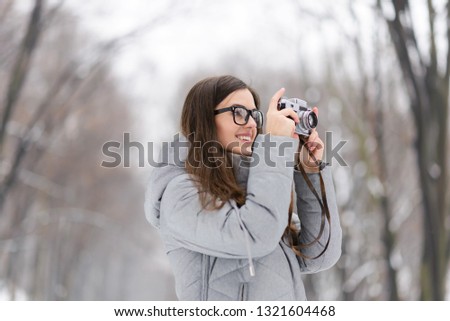 Brunette photograph girl in glasses wearing grey jacket shooting on the nature, she makes photo of winter landscapes on the vintage camera