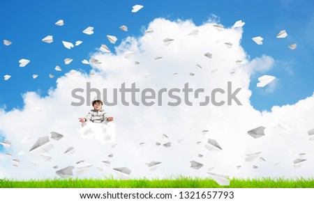 Young little boy keeping eyes closed and looking concentrated while meditating on cloud among flying paper planes with bright and beautiful landscape on background.