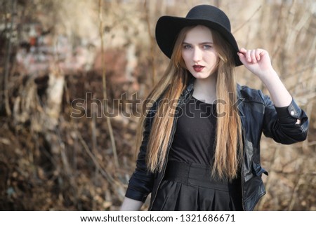 A girl walks down the city street in a leather waistcoat with a phone. Young beautiful girl in a hat and with a dark make-up outside. Girl in the Gothic style on the street.
