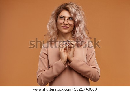 Sly hipster girl planning prank or evil trick, clasping hands and smiling mysteriously. Pensive cunning young woman in eyewear having tricky plan in mind, posing isolated at blank orange studio wall Royalty-Free Stock Photo #1321743080