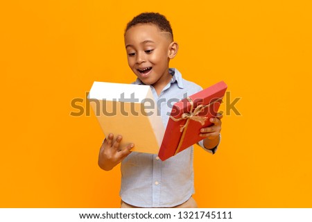 Emotional excited African American schoolboy receiving box of chocolate on his birthday, looking inside with surprised joyful facial expression, opening mouth widely, posing isolated at yellow wall