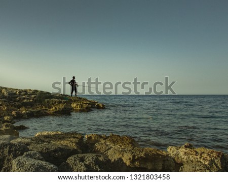 A young man catches fish in the sea from a stone shore. Silhouette of a fisherman on the horizon.